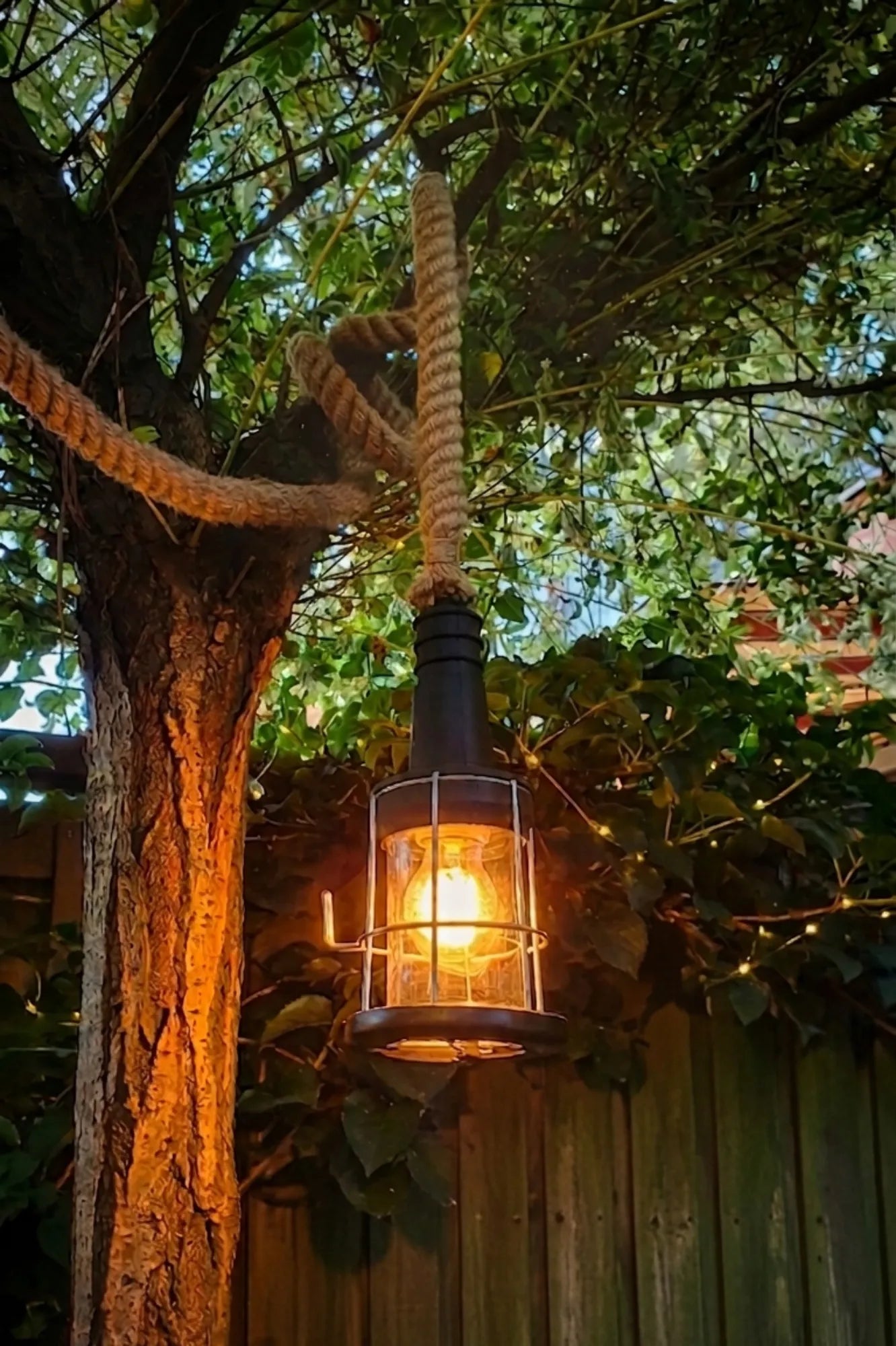 industriële buitenlamp met dik jutetouw en warme verlichting hangend aan boom in sfeervolle tuin bij schemering
