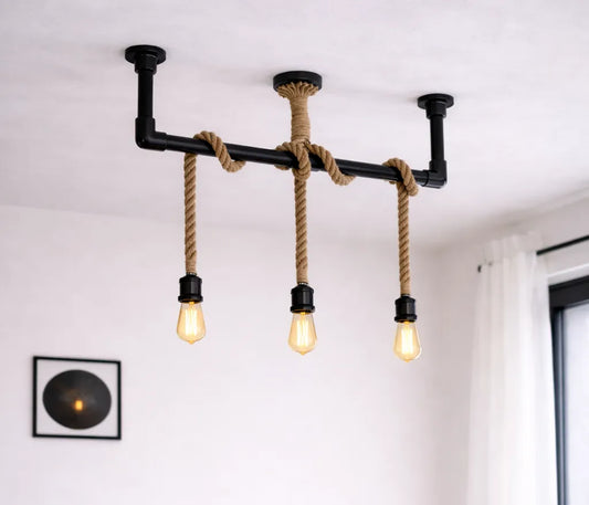 Industriële steigerbuis plafondlamp met drie touwpendels en zwarte fittingen, gemonteerd aan het plafond in een lichte moderne ruimte met witte muur en raam.