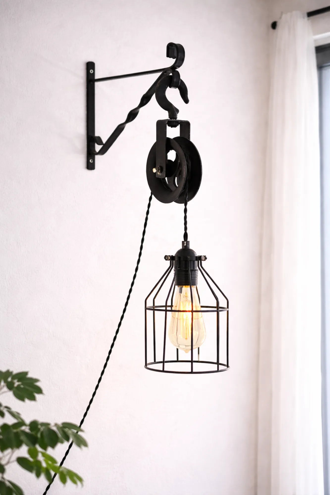 Industriële katrol wandlamp met zwarte metalen kooi en textielsnoer, gemonteerd aan de muur in een lichte woonkamer.