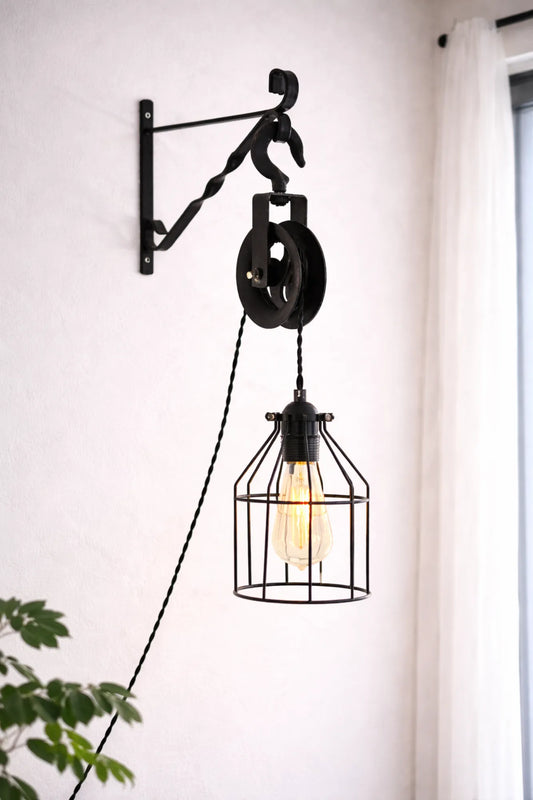 Industriële katrol wandlamp met zwarte metalen kooi en textielsnoer, gemonteerd aan de muur in een lichte woonkamer.